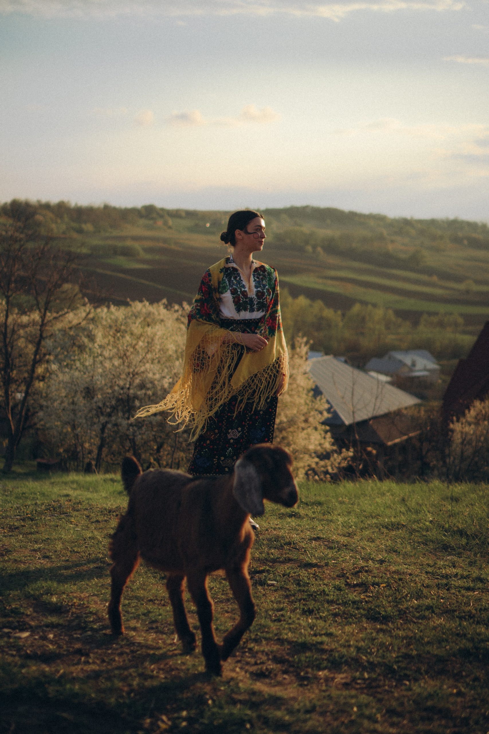 Beautifully dressed woman standing beside a goat in a mountain pasture.